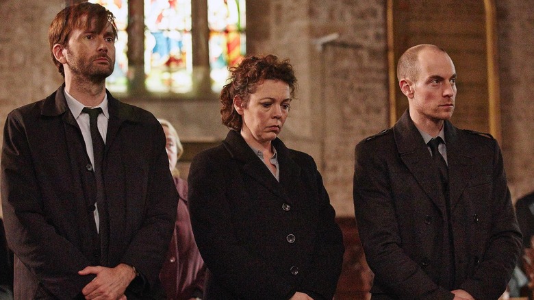 Detective Inspector Alec Hardy, Detective Sergeant Ellie Miller, and her husband, Joe Miller, standing in a church pew in "Broadchurch"