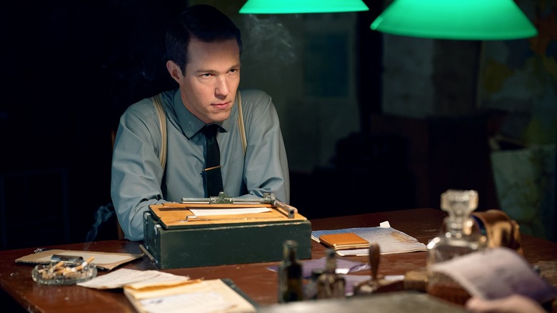 A man sits in the dark at his desk, with cigarette smoke in the air.