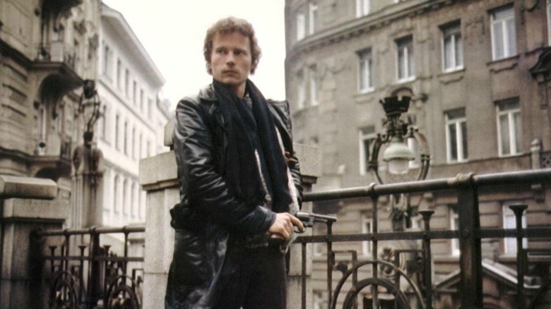 John Savage holds a gun on a rooftop