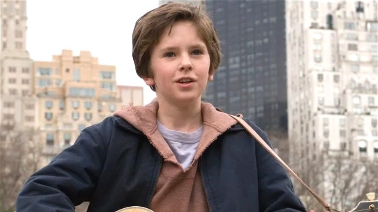 August playing a guitar outdoors with New York City buildings behind him