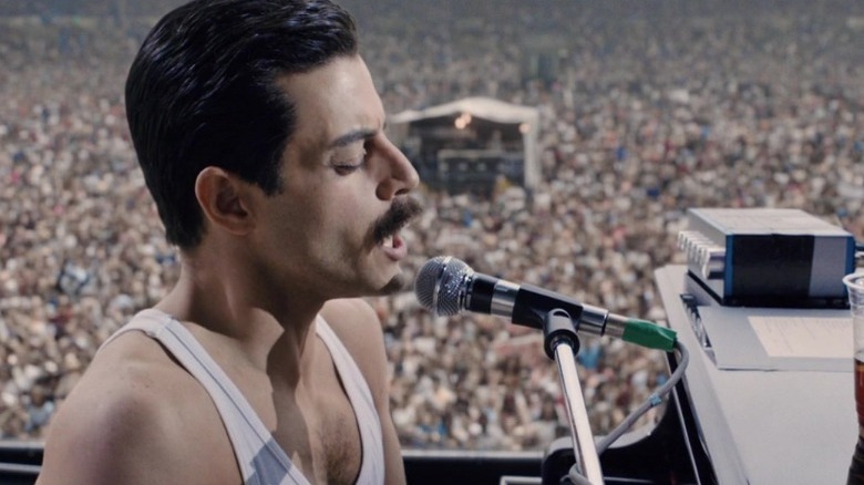 Freddie Mercury singing into a microphone at a piano with a huge crowd watching