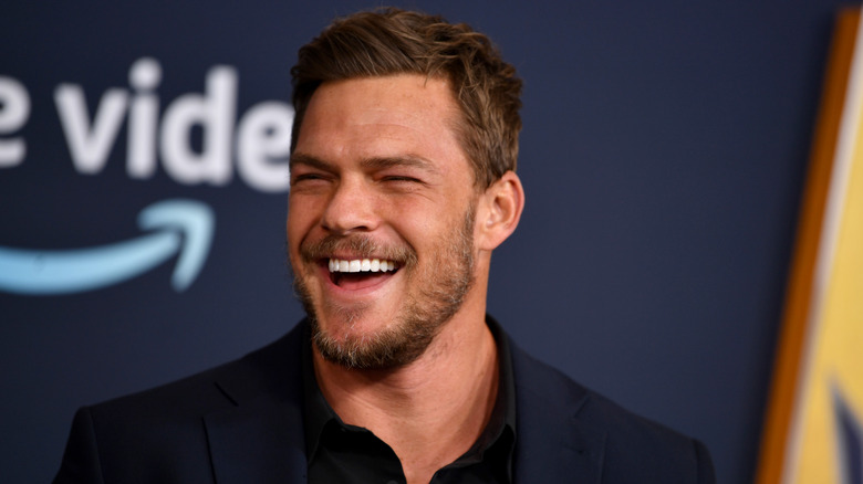 Alan Ritchson smiles in front of prime video logo