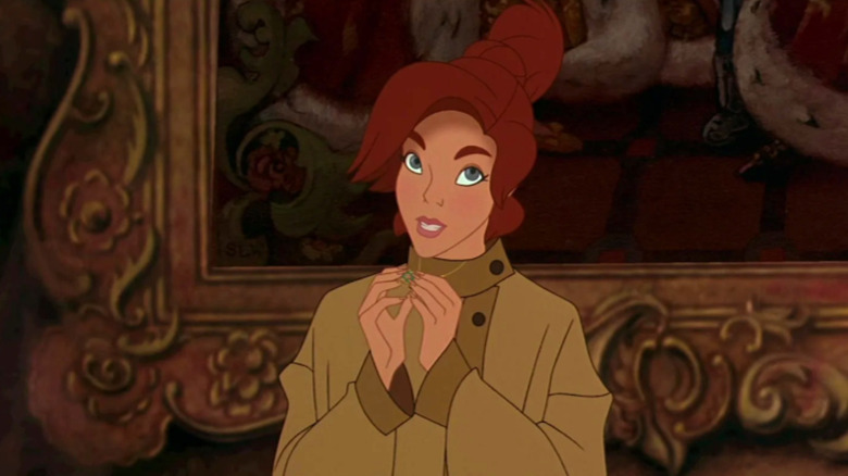 Adult Anastasia wears a beige sweater and holds her gold necklace in her hands while looking toward the right