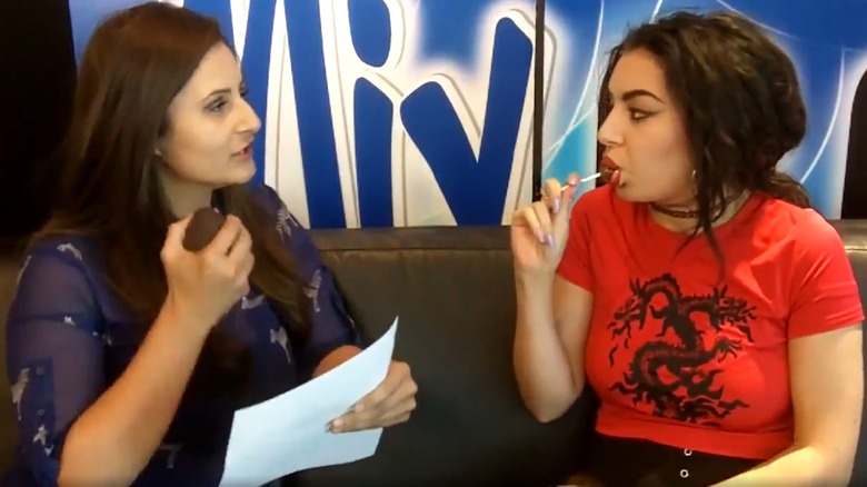 Charli XCX sitting beside a woman while sucking on a lollipop during an interview