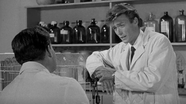 A lab assistant in a white coast stands in front of a row of bottles and talks with another man whose back is to the camera