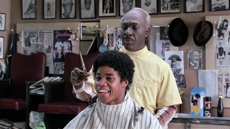 A young man smiles in a barber's chair as an elderly barber holds a scissors near his head