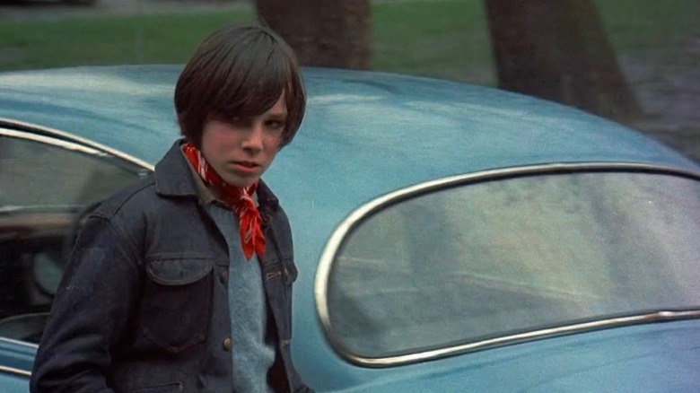 A boy stands near a car, looking suspiciously off to the side