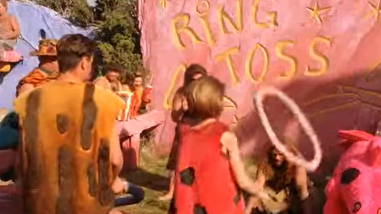 A crowd of people dressed in brightly-colored caveman outfits gather around a ring toss game