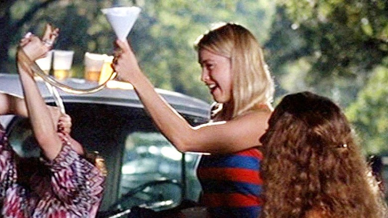 Three young women stand by a car drinking from a beer bong