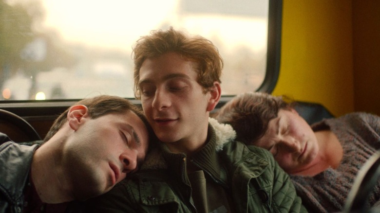 A man on a bus smiles at another man sleeping on his shoulder