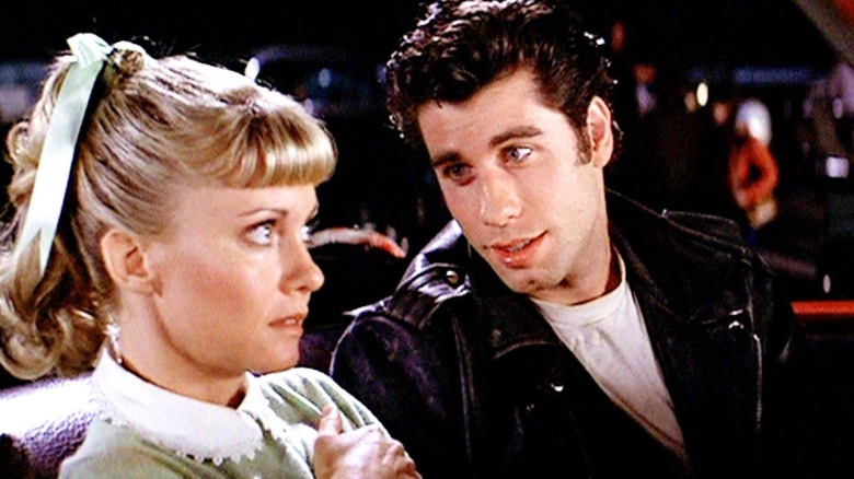 A young man in a leather jacket leans in toward a young woman in a sweater who is staring intently off-camera