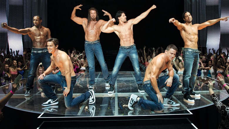 A group of six male dancers stand shirtless on a stage while a crowd around them applauds