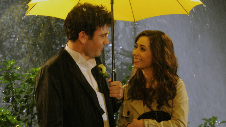 Ted Mosby and the Mother smiling at each other in the rain under a yellow umbrella in "HIMYM"