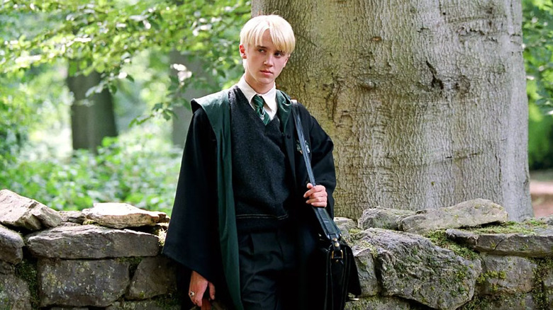 Draco leans next to a tree in Hogwarts uniform