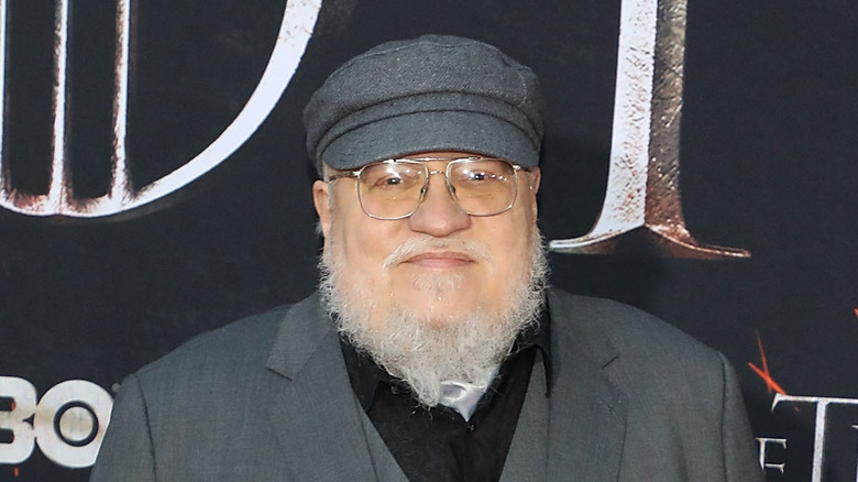 George R. R. Martin smiles in front of a promotional poster for Game of Thrones