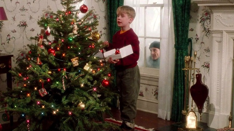 Kevin hangs ornaments on a Christmas tree as Harry looks at him through a window