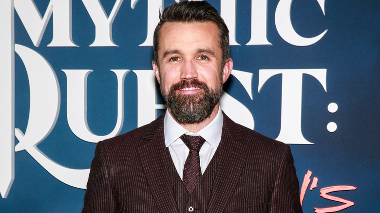 Rob McElhenney smiling in front of a Mythic Quest display 