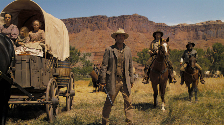 Old cowboy holding rifle near cowboys on horses and caravan of family