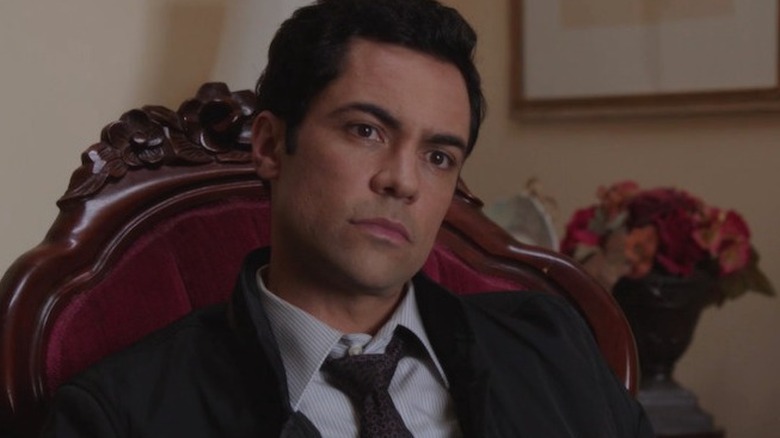 Detective Amaro sitting in velvet and wood chair 