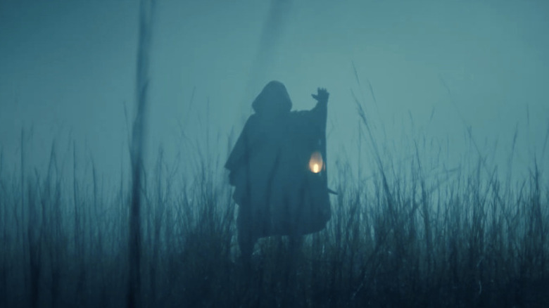 Hooded figure in a field holding a lamp in Loki