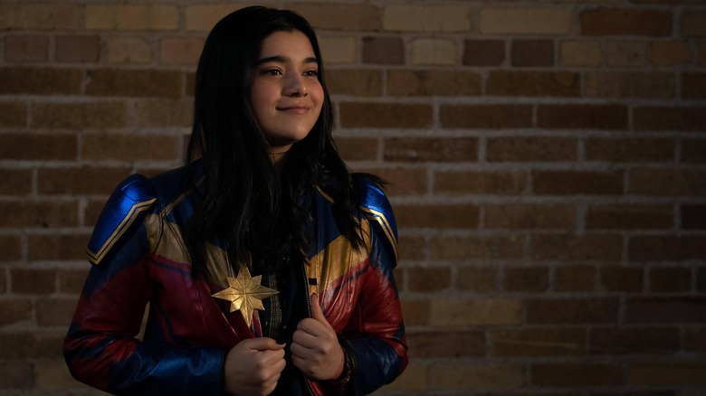 Ms. Marvel smiling in Captain Marvel jacket next to brick wall