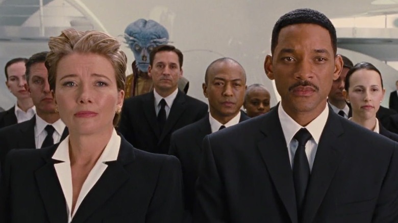 Agent O and Agent J stand at the front of an assembly of Men in Black agents wearing suits