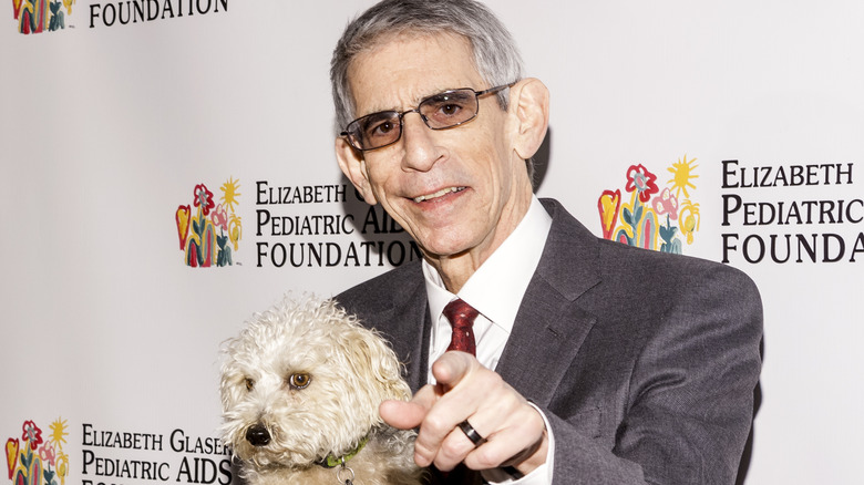 Richard Belzer holding a dog and pointing at the camera