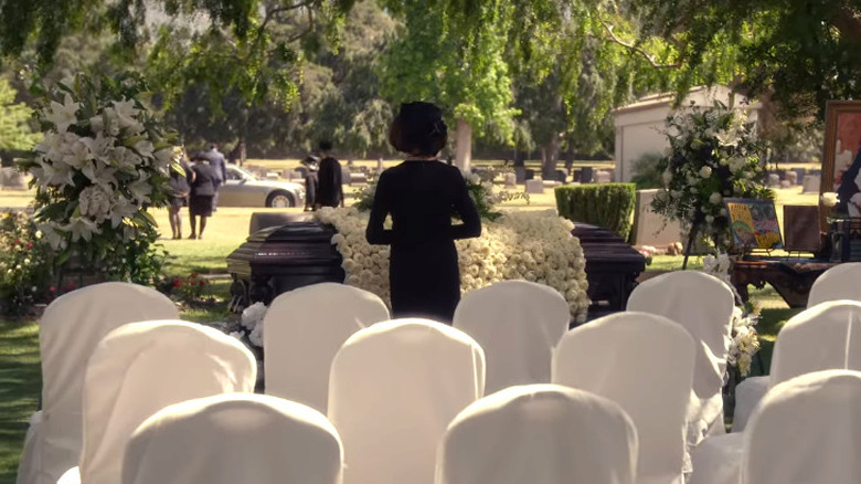 Emily stands in front of Richard's casket draped with white flowers in the graveyard