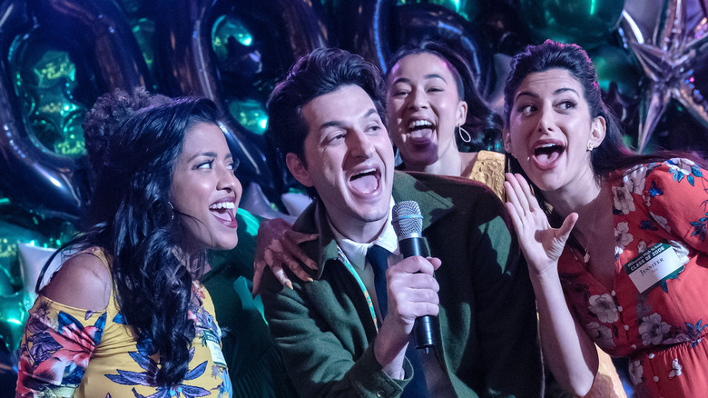 Ben Schwartz sings into a microphone surrounded by backup singers in The Afterparty