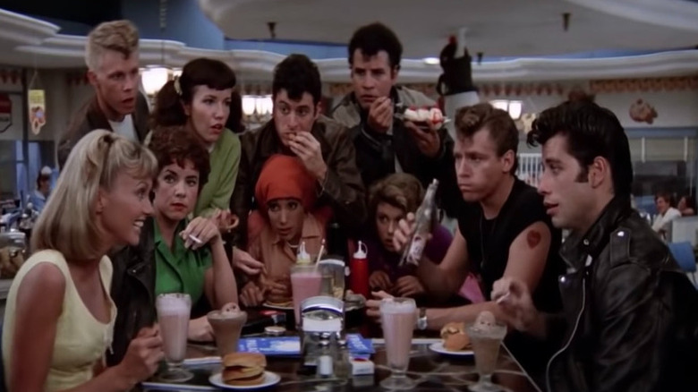 Sandy and Danny sit at their diner table while the Pink Ladies and the T-Birds gather around them