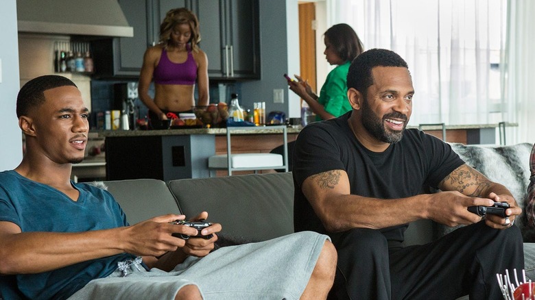 Two men play video games while two women work in the kitchen