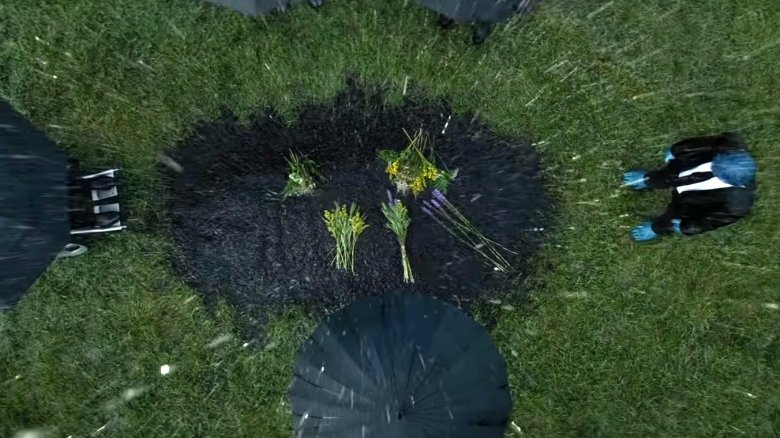 An overhead shot of a freshly dug grave covered in flowers and surrounded by mourners in the rain