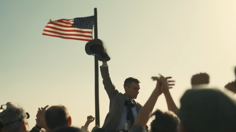 Oppenheimer waving to applauding crowd next to American flag