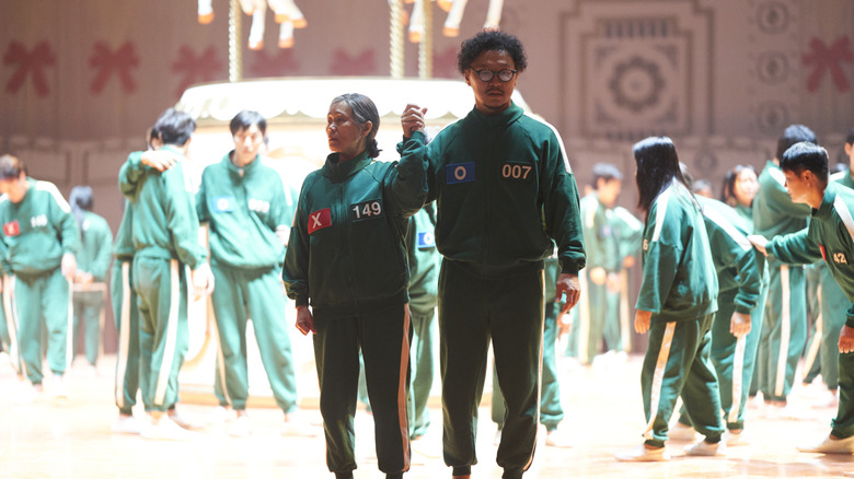 Kang Ae-shim as Jang Geum-ja, player 149, and Yang Dong-geun as Park Yong-sik, player 007, holding hands in the middle of the game Mingle on Squid Game Season 2