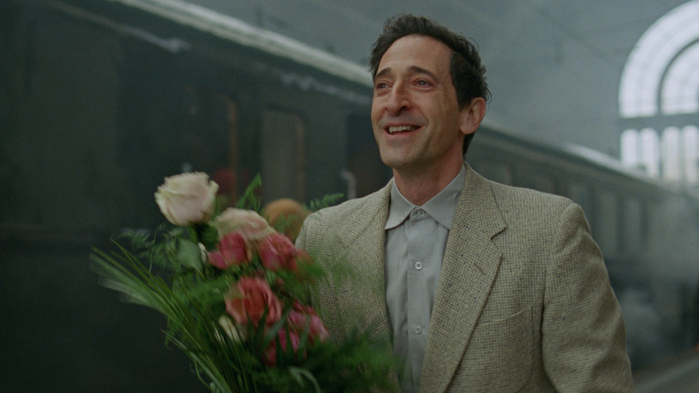 László Tóth holding flowers and smiling at a train station in The Brutalist