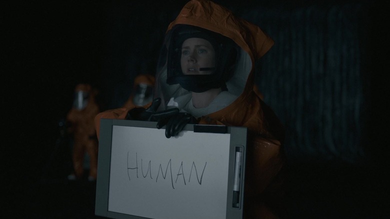 Louise, wearing a hazmat suit, holds a paper reading "human."