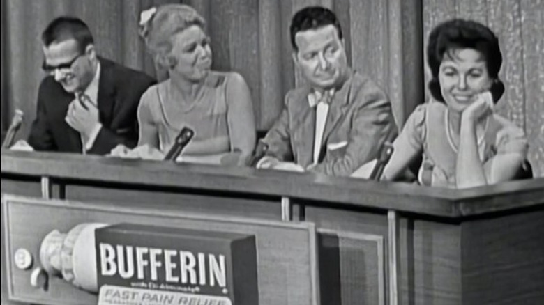 Four people behind a game show counter and microphones in black and white