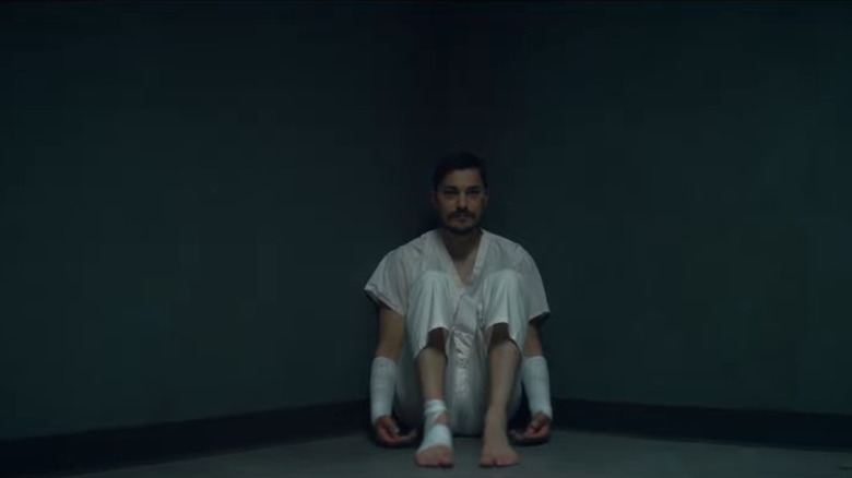 Man in white clothes and bandages sitting in corner of room 