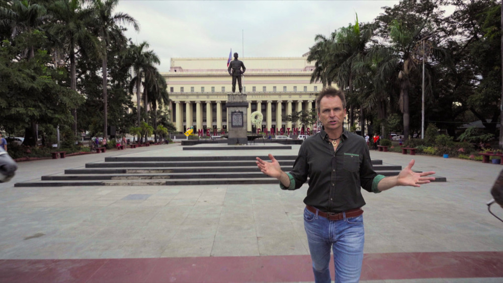 Phil Keoghan stands in front of statue 