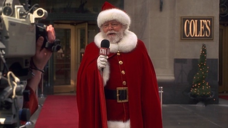 Richard Attenborough as Kris Kringle holding a microphone and addressing a camera while dressed as Santa Claus in Miracle on 34th Street (1994)