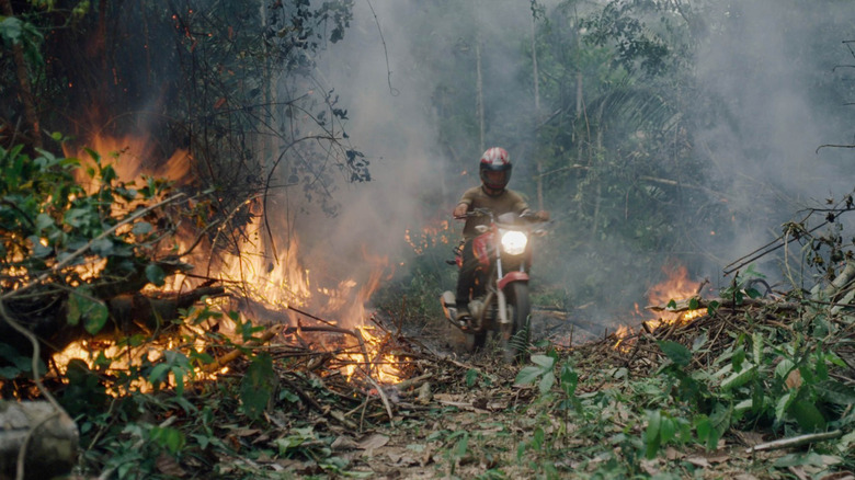 A figure rides a motorcycle through a burning forest