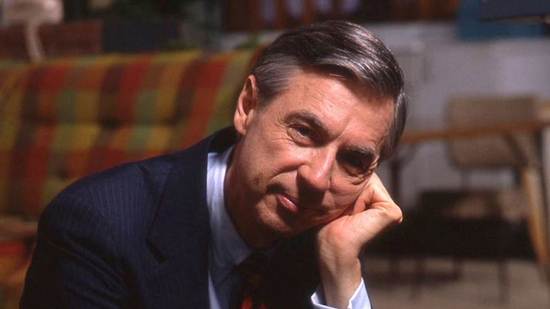 Mister Rogers sits with his hand supporting his head and a small smile on his face