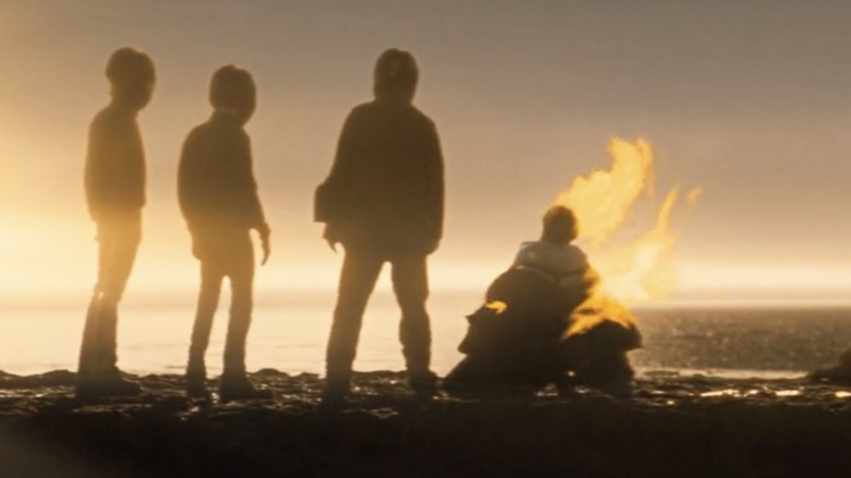 Three silhouettes stand on a beach at sunset, with another on the ground on fire