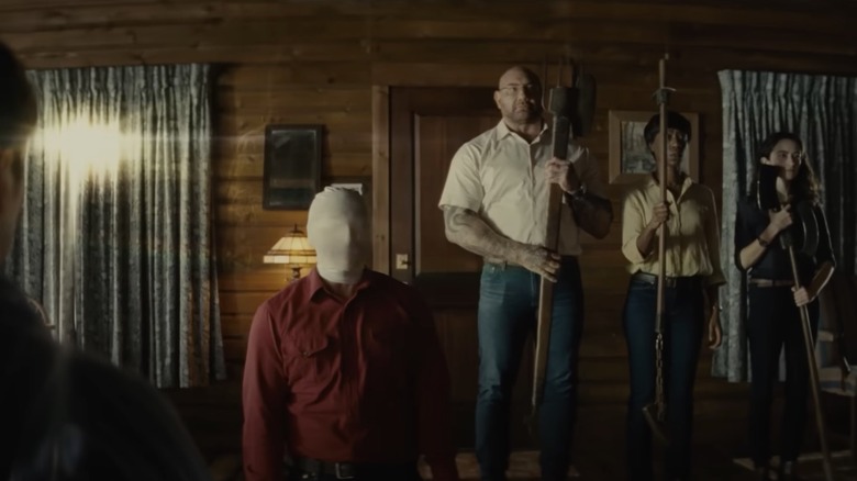 Three strangers stand with weapons behind man kneeling in mask