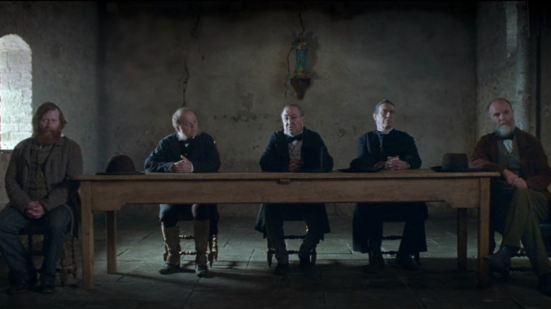 Five men sit in a row at a table in a sparse room