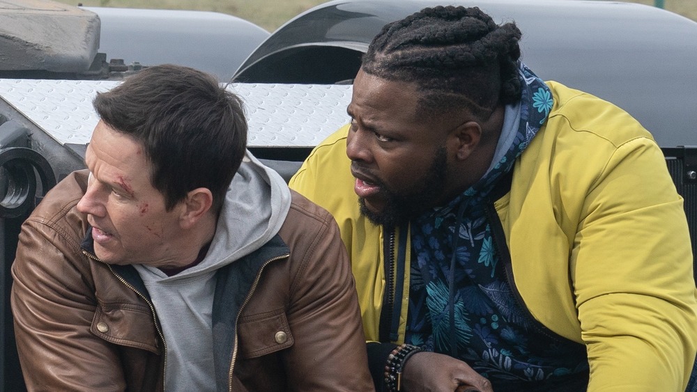 Mark Wahlberg and Winston Duke crouched behind a car in Spenser Confidential