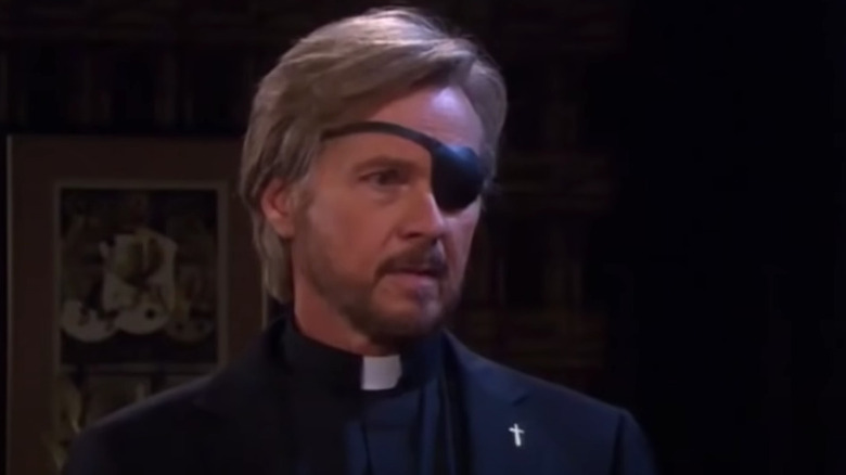 A priest in an eyepatch stands in an office 