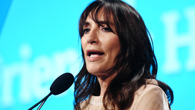 Katey Sagal speaking into a microphone, turned to the left, with a light blue background