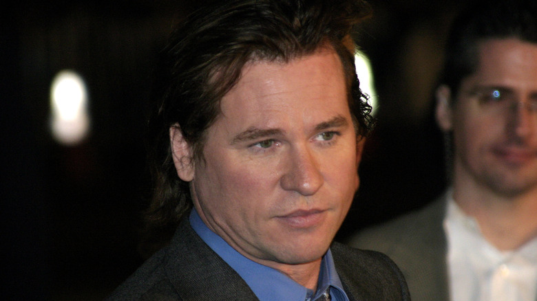 Val Kilmer looking serious to the right, wearing a blue shirt and grey jacket, at an event in 2004