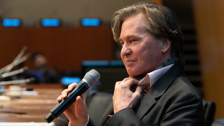 Val Kilmer at a 2019 event facing left, holding a microphone, with his hand on his shirt collar